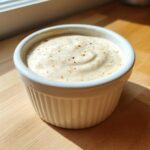A small white ramekin filled with creamy Hot Honey Ranch Sauce, sprinkled with spices, sitting on a wooden counter.