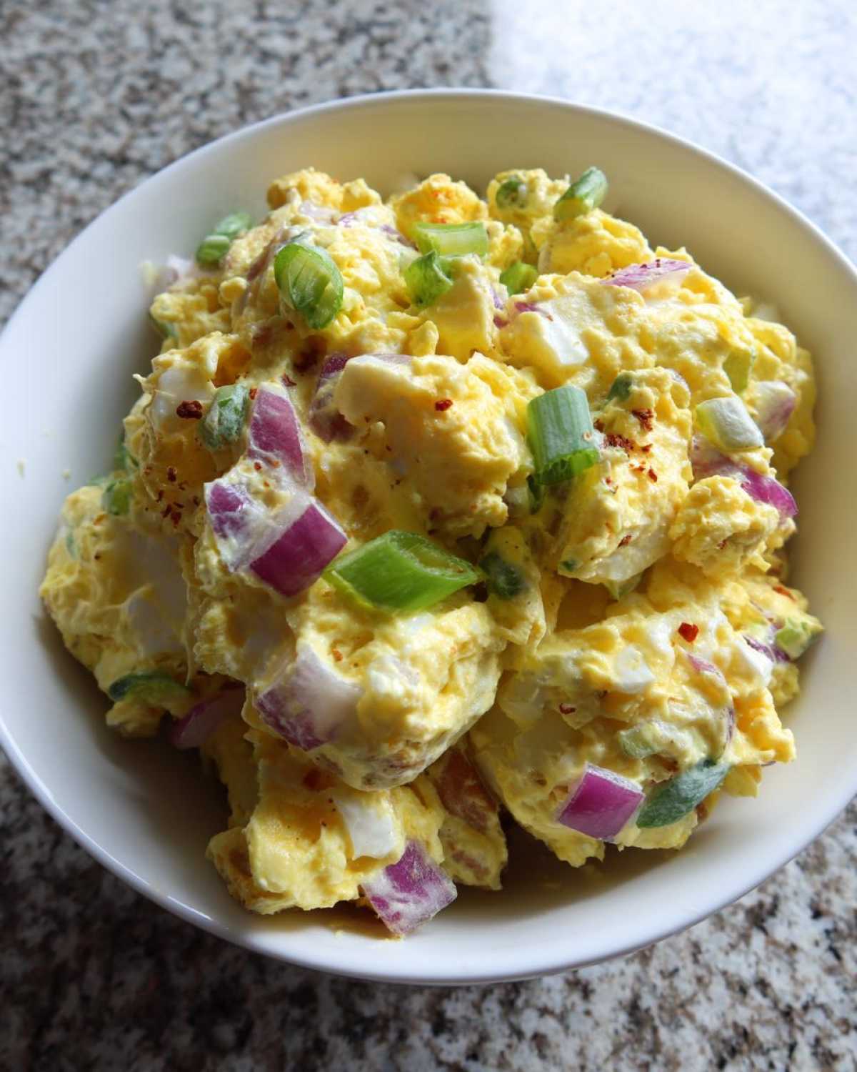 Close-up of creamy Jamaican Jerk Potato Salad mixed with chunks of red onion and green onions in a white bowl.