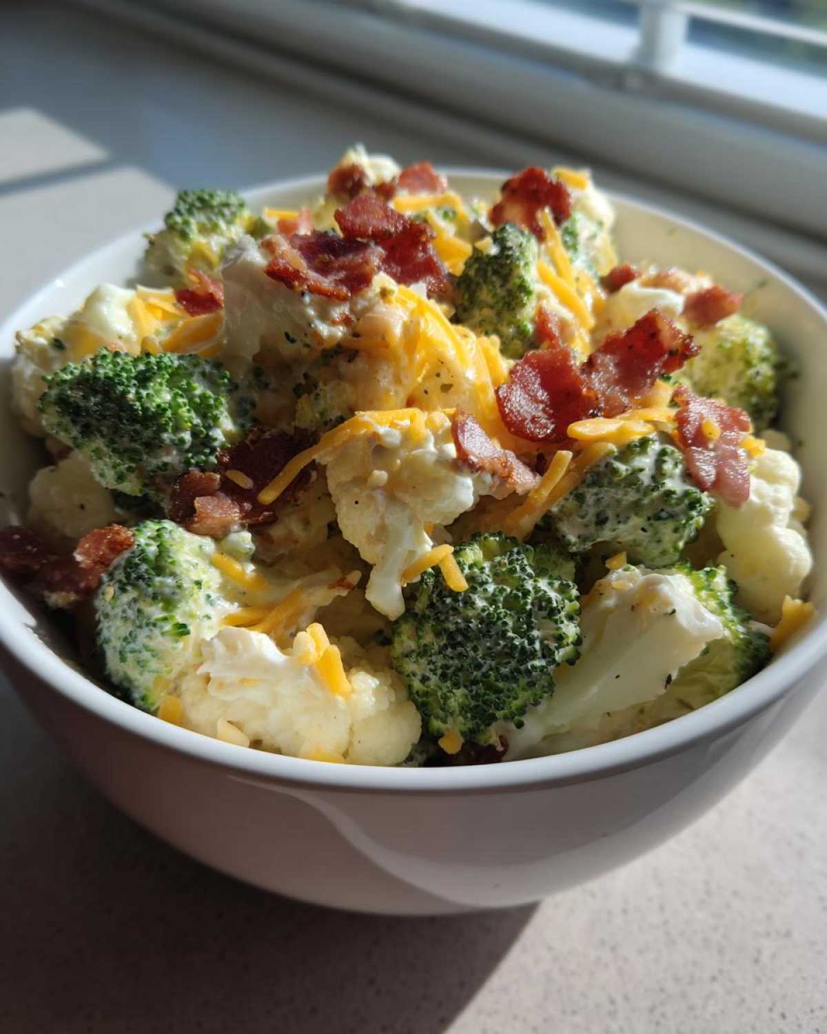 A white bowl filled with Loaded Broccoli Cauliflower Salad Low Carb Incredible, topped with bacon bits and shredded cheese.