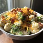 A close-up of the Loaded Broccoli Cauliflower Salad Low Carb Incredible, topped with shredded cheddar cheese and bacon bits in a white bowl.