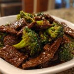 A close-up serving of glossy, saucy Low Carb Mongolian Beef mixed with bright green broccoli florets in a white bowl.
