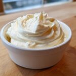 A close-up of thick, creamy, pale yellow Lowcarb Garlic Aioli swirled high in a small white ramekin on a wooden surface.