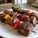 Close-up of juicy, grilled Steak Kabobs featuring seared beef, red onion, bell peppers, and mushrooms on a white plate.