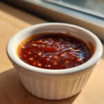 Close-up of rich, dark Pineapple Jalapeno Bourbon BBQ Sauce with visible chili flakes in a small white ramekin.
