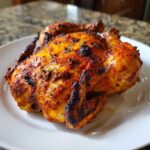 A whole, perfectly roasted Pollo Asado with crispy, charred, reddish-orange skin resting on a white plate.