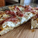 A close-up view of a slice of Prosciutto Honey Burrata Pizza showing creamy burrata, draped prosciutto, and a drizzle of honey on a wooden board.