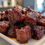 Close-up of glistening, dark brown Sticky Steak Bites piled on a white rectangular plate.