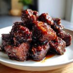A close-up of a pile of glistening Sticky Steak Bites coated in a dark, thick glaze served on a white plate.