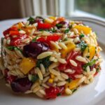 A molded serving of Veggie Rainbow Orzo Salad featuring orzo pasta, colorful bell peppers, and Kalamata olives.